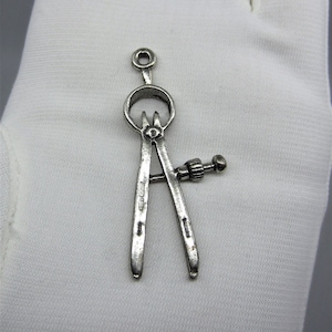 Può includere: Un ciondolo a forma di bussola in argento con un design dettagliato. La bussola ha una base circolare e due bracci che si incontrano in un punto. Il ciondolo è su una superficie bianca.