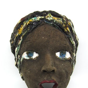 Può includere: Una maschera in ceramica con un viso marrone scuro, occhi blu e labbra rosse. La maschera è decorata con un motivo colorato intorno alla testa.