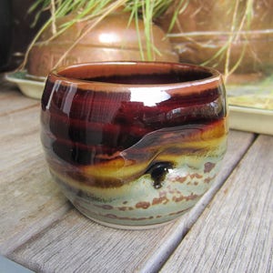 May include: A small, brown, glazed ceramic cup with a speckled design. The cup has a rounded bottom and a wide rim.