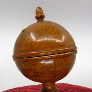 Könnte beinhalten: Eine hölzerne, kugelförmige Box mit einem Scharnierdeckel und einem kleinen Knopf oben. Die Box steht auf einem Holzsockel und hat eine satte braune Farbe.