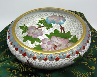 May include: A white cloisonné bowl with a gold rim and floral design. The bowl is decorated with pink and green flowers and leaves on a white background. The bowl is sitting on a green fabric with a gold pattern.