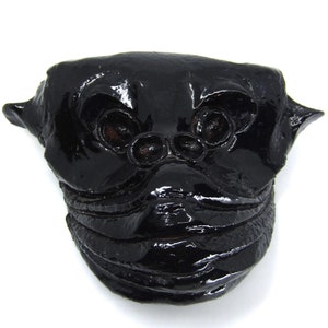 Könnte beinhalten: Eine schwarze Keramikmaske mit groteskem Ausdruck. Die Maske hat große Augen, einen breiten Mund und eine strukturierte Oberfläche.