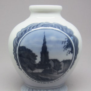 Peut inclure: Un vase en céramique blanche avec un motif bleu et blanc. Le vase représente une scène d'église avec un haut clocher et des bâtiments environnants. Le motif est encadré par une bordure décorative.