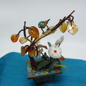 Puede incluir: Un bonsái de jade en miniatura con un pájaro posado en una rama. El árbol está hecho de jade tallado y tiene una maceta pequeña con un acabado de esmalte azul y blanco.