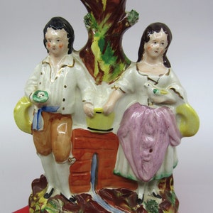 Op de afbeelding: Een vintage keramieken beeldje met een man en een vrouw aan weerszijden van een boomachtige structuur met een rode binnenkant. De figuren dragen kleding uit die tijd en de basis is versierd met bladeren en een rode bakstenen structuur.