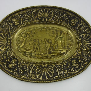 May include: An oval brass tray with an embossed design. The center of the tray features a scene of three figures in a garden setting. The border of the tray is decorated with a floral and shell pattern.
