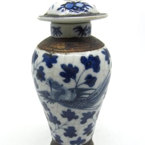 May include: A small, antique ceramic vase with a lid. The vase is white with blue floral and bird designs. The lid has a small, white figurine on top. The base and the area below the lid are brown.