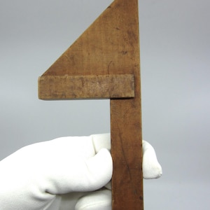 Może przedstawiać: Zabytkowy drewniany kątownik, trzymany przez dłoń w rękawicy. Narzędzie posiada trójkątne ostrze i prostopadły uchwyt, oba wykonane ze starego, barwionego drewna. Otwór jest widoczny w pobliżu końca uchwytu. Jest to narzędzie stolarskie.