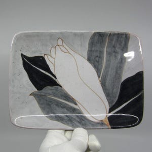 May include: A white ceramic rectangular dish with a black and gray floral design. The design features a white flower with gray leaves.