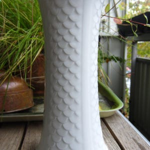 May include: White ceramic vase with a textured, scale-like pattern. The vase is sitting on a wooden table.
