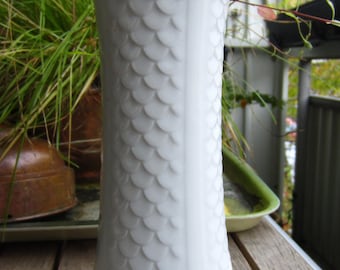 May include: White ceramic vase with a textured, scale-like pattern. The vase is sitting on a wooden table.