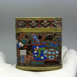 Peut inclure: Une petite boîte rectangulaire en laiton avec un couvercle à charnière. La boîte est décorée de cloisonné en émail coloré, avec un motif floral aux accents bleus, rouges, verts et blancs.