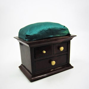 Può includere: Un piccolo forziere di legno con tre cassetti e un cuscino di velluto verde in cima. Il forziere ha pomelli dorati sui cassetti.