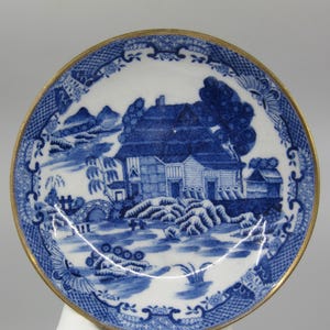 May include: A blue and white porcelain saucer with a gold rim. The saucer features a traditional Chinese landscape scene with a house, trees, and a bridge.