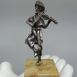 May include: Silver sculpture of a person playing a violin, standing on a beige marble base with a silver plaque that reads "A. Kedem Israel".