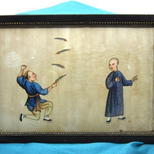 Op de afbeelding: Een antiek ingelijst schilderij met twee figuren in traditionele kleding. Eén figuur gooit messen, de ander staat ernaast. Het schilderij heeft een zwarte lijst met een kralenrand en een crèmekleurige achtergrond.