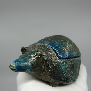 Op de afbeelding: Een keramieken egelbeeldje met een scharnierend deksel, met een getextureerd oppervlak en een mix van blauwe, groene en gouden tinten. Deze kleine decoratieve doos is een uniek woonaccessoire.