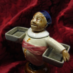 Puede incluir: Una figurita de cerámica de una persona que lleva un sombrero azul y una camisa blanca con una faja rosa. La figurita sostiene dos bandejas en sus manos.