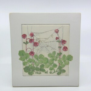 May include: A square ceramic tile featuring a raised relief design of red flowers and green leaves growing in front of a spider web. The background of the central panel is off-white, framed by a smooth, light grey border with a matte finish.