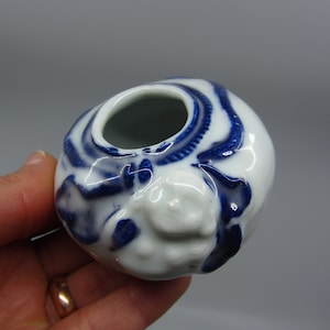 Puede incluir: Una pequeña maceta de cerámica blanca con diseños florales azules y blancos. La maceta tiene forma redonda y una pequeña abertura en la parte superior.