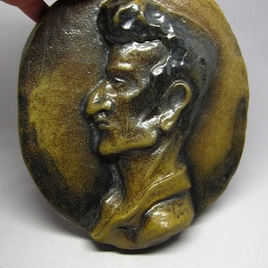 Puede incluir: Una placa redonda de cerámica marrón con una escultura en relieve de un perfil de hombre. El hombre tiene el pelo corto y oscuro y lleva una camisa.