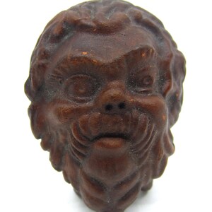 May include: A detailed, brown, carved wooden face with a whimsical expression. The face has a prominent nose, a slight smile, and detailed hair. The carving is set against a white background.