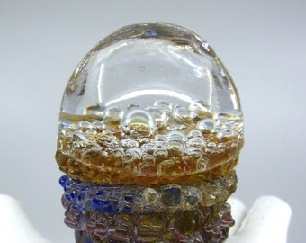 May include: A clear glass paperweight with a layered design. The top layer is clear glass with air bubbles. The middle layer is a light brown color. The bottom layer is a mix of blue, purple, and gold glass.