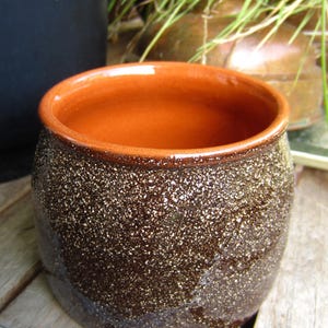 Puede incluir: Una maceta de cerámica marrón con un esmalte moteado y un borde de color terracota. La maceta está sentada sobre una superficie de madera.