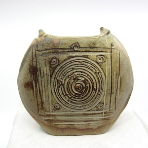 May include: A ceramic vase with a light brown glaze and a geometric design. The vase has a square shape with a circular design in the center.