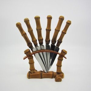 May include: A set of six silver knives with bamboo handles, displayed in a bamboo stand. The stand has a curved top and is made of natural bamboo.
