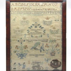 May include: A framed antique sampler with a detailed needlework design. The sampler features embroidered letters, numbers, floral motifs, and a bird. The text includes names and dates, all within a dark wood frame.