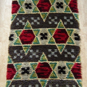 May include: A brown, green, white, and red geometric patterned rug with a shaggy texture. The rug has a repeating pattern of stars and triangles.