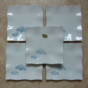 May include: Set of five white ceramic square plates with a blue floral design. The plates have a slightly wavy edge and are decorated with a stylized iris flower and leaves. One plate has a gold label with the text "寿田屋" in Japanese.