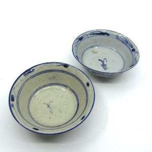 May include: Two small, off-white ceramic bowls with blue painted rims and interior designs. The bowls feature a simple, elegant design, with a delicate blue floral pattern around the edges and a small blue symbol in the centre.