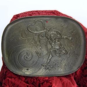 May include: A gray metal tray with a raised design of a mythical creature with a long tail and a swirling wave pattern. The tray is oval shaped and has a slightly raised rim.