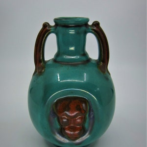 Puede incluir: Un jarrón de cerámica de color verde azulado con dos asas y un diseño en relieve de una cara en marrón rojizo en la parte delantera.