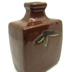 May include: A square-shaped ceramic vase with a glossy brown glaze. The vase features a narrow neck and a small, dark, abstract brushstroke design on the front surface. The body of the vase is flat and rectangular with rounded edges.