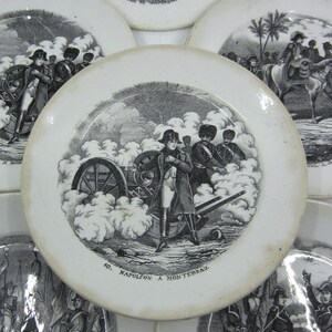 May include: Six vintage ceramic plates with black and white illustrations of military scenes. The plates feature scenes of Napoleon Bonaparte and his troops in battle. The plates are decorated with a black and white design and have a white background.