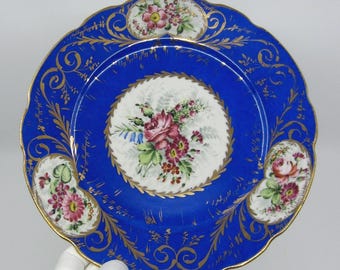 May include: A round porcelain plate featuring a deep blue border with intricate gold filigree patterns. The center and three scalloped edge panels display hand-painted floral bouquets in shades of pink, red, green, and white on a white background.