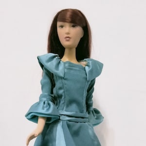 Puede incluir: Un vestido azul turquesa con cuello y mangas con volantes. El vestido tiene un corpiño ajustado y una falda completa. La muñeca lleva el vestido y tiene el pelo castaño y los ojos azules.
