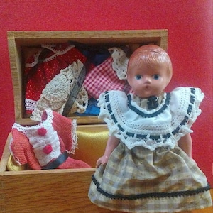May include: A porcelain doll with brown hair and blue eyes wearing a white and black lace collar and a plaid dress with black trim. The doll is sitting in a wooden box with a variety of fabric scraps and lace.
