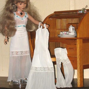 Könnte beinhalten: Eine Puppe trägt ein weißes Kleid mit einem durchsichtigen, plissierten Rock und floraler Spitzenborte. Die Puppe hält ein passendes weißes Kleid. Ein Holzschreibtisch mit einem Teeservice befindet sich im Hintergrund.