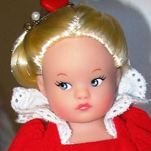 May include: A doll with blonde hair styled in a bun, blue eyes, and rosy cheeks. The doll wears a red dress with a white lace collar and a red heart-shaped hair accessory. A pearl and silver-colored metal adorn the doll's hair.