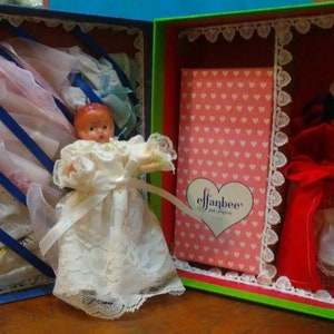 Puede incluir: Una casa de muñecas vintage con dos habitaciones, una con una muñeca vestida con un vestido blanco y la otra con una muñeca vestida con un vestido rojo y blanco. La casa de muñecas está decorada con encaje y corazones.