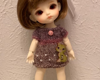 May include: A doll with brown hair and red eyes wearing a knitted purple dress with a green cat applique and pearl accents. The doll is wearing yellow shoes with white bows.