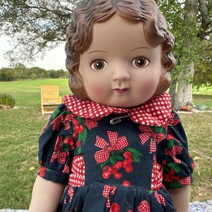 May include: A vintage doll with brown hair and eyes, wearing a navy blue dress with a red cherry and bow pattern. The dress has a red and white checkered collar and is secured with a safety pin. The doll is posed outdoors.