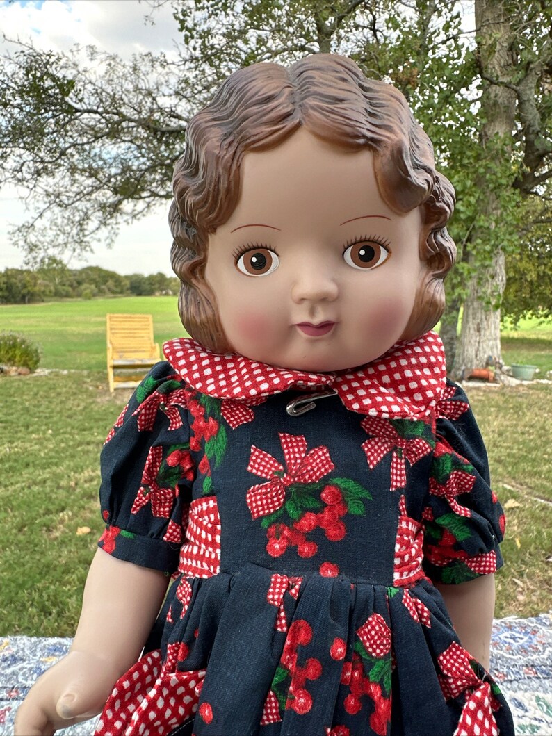 May include: A vintage doll with brown hair and eyes, wearing a navy blue dress with a red cherry and bow pattern. The dress has a red and white checkered collar and is secured with a safety pin. The doll is posed outdoors.