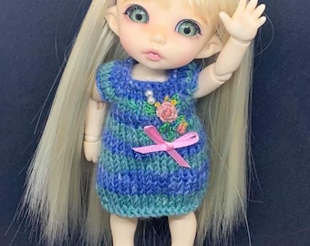 May include: A doll with long blonde hair and green eyes, wearing a blue and green knit dress with a pink bow and floral embroidery. The doll is also wearing pink shoes with a cat face design.