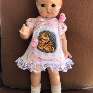 May include: A vintage doll wearing a pink knitted dress with a cat embroidery and lace trim. The doll has a pink headband and white shoes. Two pink bird ornaments are attached to the dress.