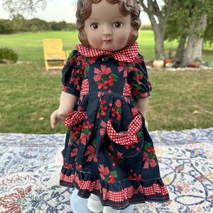 May include: A vintage doll with brown hair and eyes, wearing a navy blue dress adorned with red cherries and bows. The dress features a red and white checkered collar, pockets, and trim. The doll is wearing white shoes.